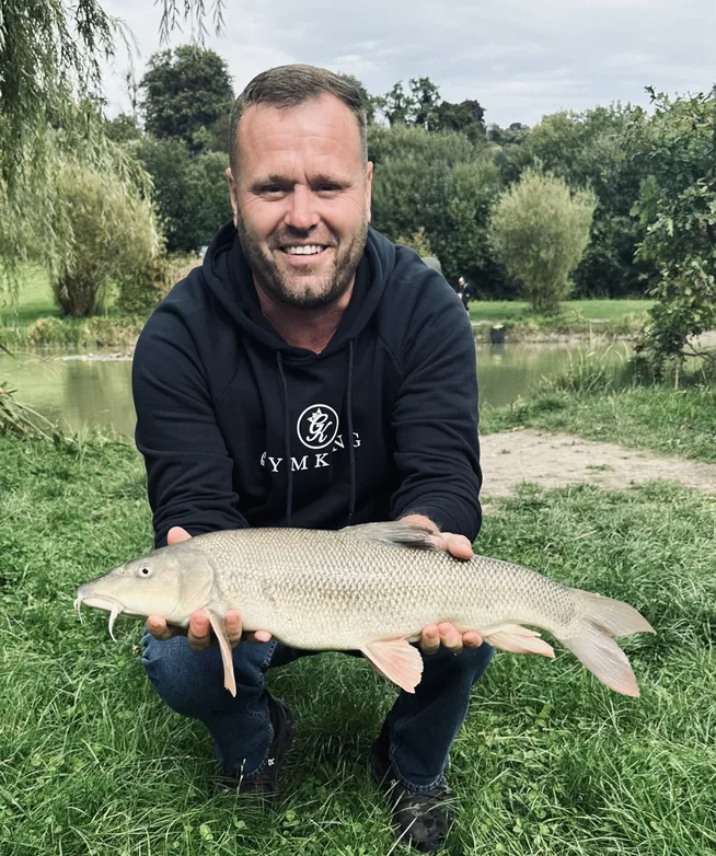 Barbel-Ben-Optimised Angler holding a barbel beside a fishing lake